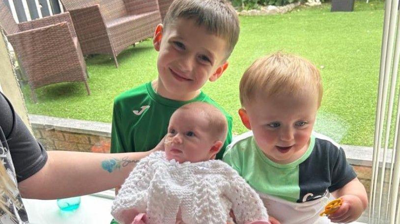 Baby Maeva is in the middle of the of her two brothers. She is wearing a white knitted jumper and is being held by her mum, who is out of shot. Her brothers are wearing green T-shirts. A garden can be seen through a window behind them.