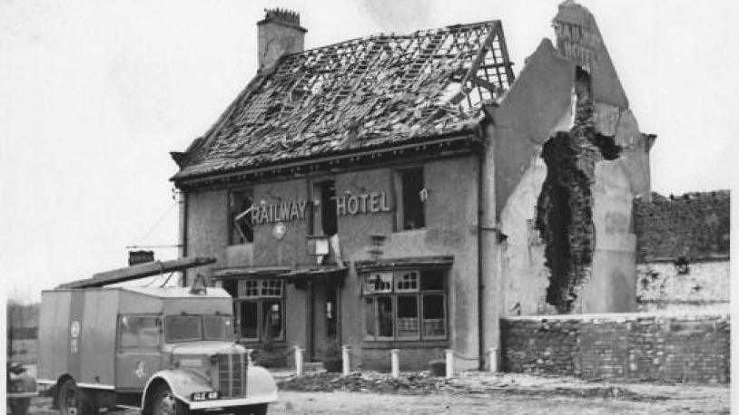 An exterior shot of the former Railway Hotel in Catterick, North Yorkshire, after an ammunition explosion in 1944. The roof is destroyed and there is a large hole on the right hand side wall. Parked outside is a fire engine. 