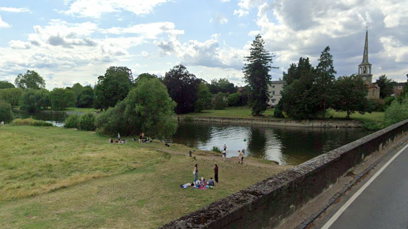 The River Thames at Wallingford, a grassy bank by the river where people are sat on blankets. Some are in the water. In the background is St Peter's Church, the tallest building in view, which has a pointed spire.