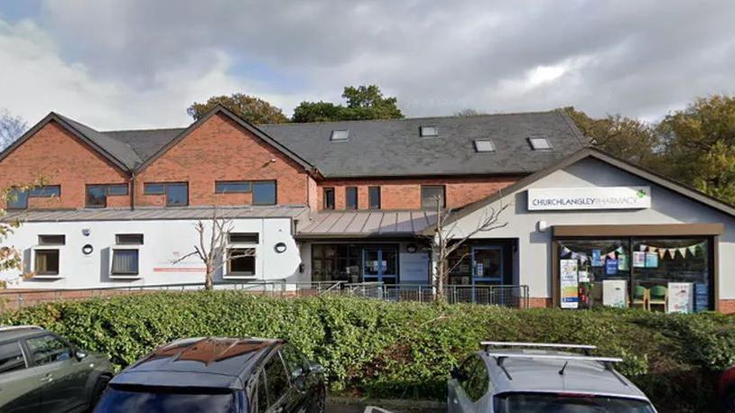 A view of a building housing a medical practice with a pharmacy attached to it. The building is mainly red brick with white rendering on the ground floor level. There are cars in a car park in front of the building