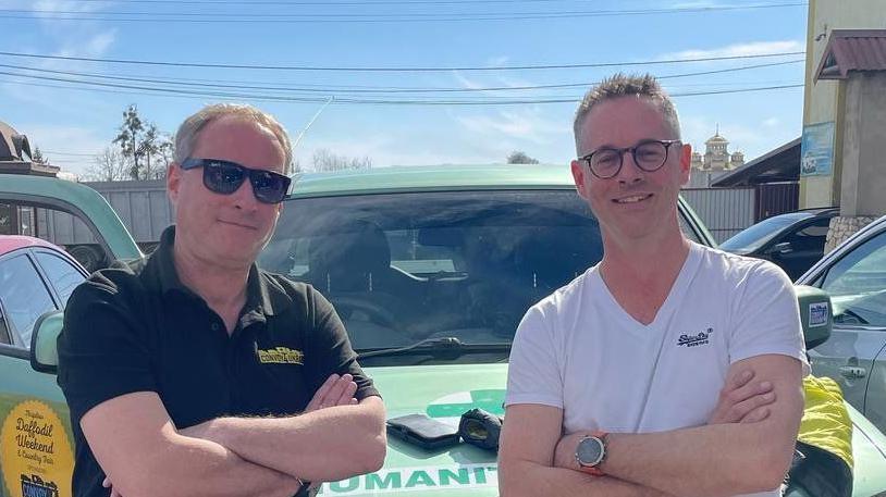 Paul Earnshaw and Alastair Gray are standing in front of the green jeep with their arms folded over. Paul is wearing a black top and black sunglasses. Alastair  is wearing a white top and glasses. 