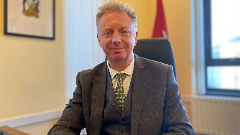 Alex Allinson sitting at his office desk with a standing Isle of Man flag in the background. He has short, fair, wavy hair and is wearing a grey suit and waistcoat with a green patterned tie and white shirt.