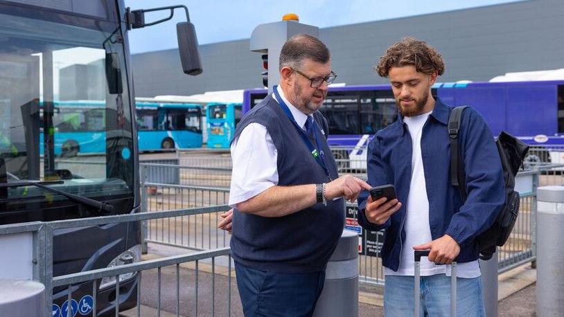 Two people stand beside a large bus outside London Luton Airport. One person is showing something on a smartphone screen while the other person, who is carrying a backpack and holding the handle of a wheeled suitcase, looks at the device.