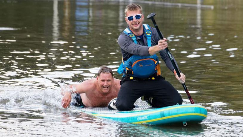 Two men on a paddleboard, the one in front is kneeling and has shades and the paddle. The one behind is lying down on the board and is topless.