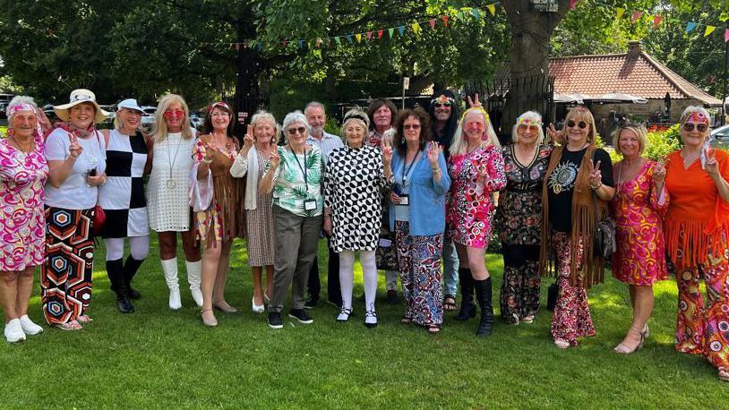 The Washington Bloom Group on the village green dressed in colourful 1960s costumes. There are trees and bunting in the background. They are smiling and many of them are doing a peace gesture. 