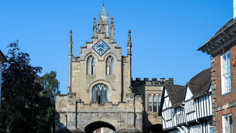 A grey/brown chapel style building with spires and arches windows, and a blue and gold clock. It sits on top of a gatehouse with an archway. it is bordered by black and white houses on its right and trees on its left