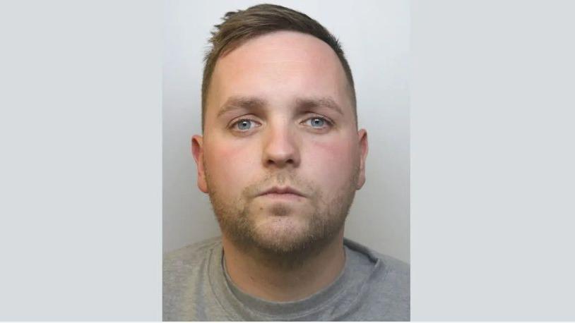 Sam Jones looks into the camera. He is not smiling. He wears a grey jumper and has a white wall behind him. He has brown hair and blue eyes.