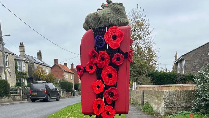 A postbox topped with knitted soldiers boots attached to poppies cascading down either side. Village street behind.