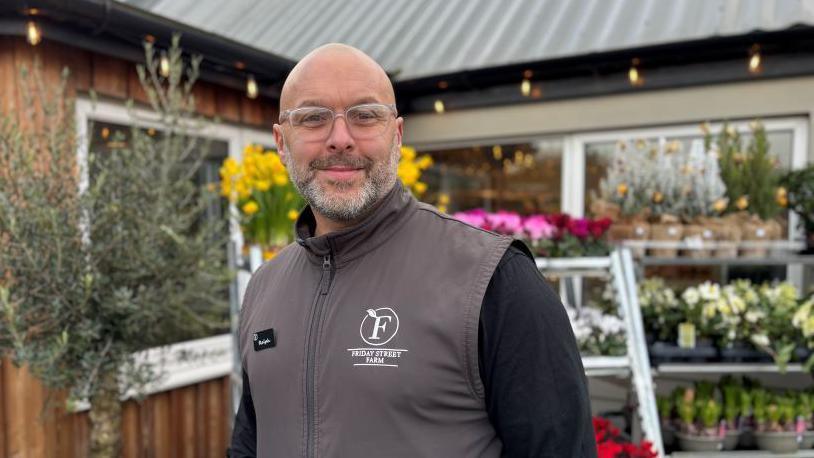 Ralph Bishop smiles into the camera. He is bald and has a grey beard and is wearing glasses. Behind him are shelves stocking flowers and plants.