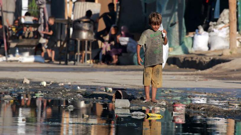 A Palestinian boy stands barefoot near stagnant wastewater in Deir el-Balah, in the central Gaza Strip (19 July 2024)