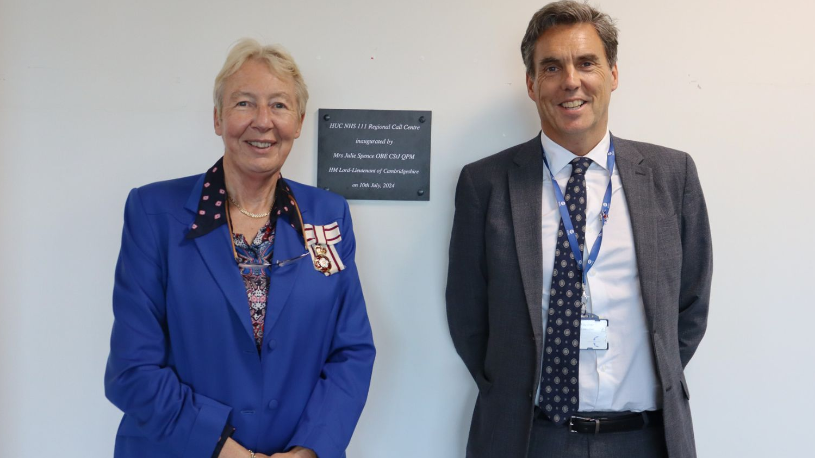 Julie Spence and David Archer stand by a plaque placed on a white wall. Julie is wearing a blue jacket and David a grey jacket, along with a shirt and tie.