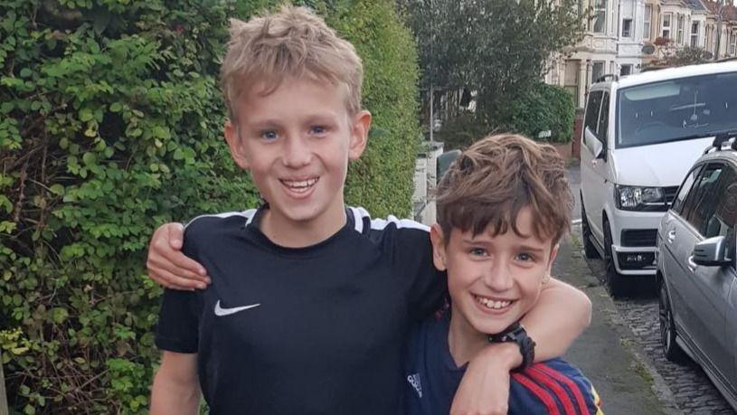 George (left) and Artie (right) smile at the camera with their arms around each other's shoulders. George has blonde hair and wears a black Nike t-shirt, while Artie has brown hair and wears a red and blue striped football shirt.