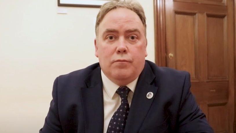 A middle aged man sitting down in an office - he is wearing a dark blue suit, a white shirt and blue patterned tie. A wooden door is visible behind him.
