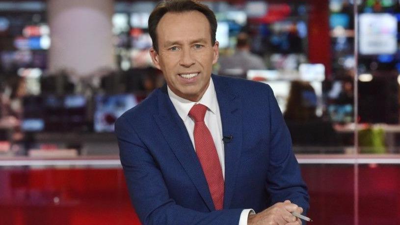 Ben Brown sits behind a desk in the One O'Clock News studio. He is wearing a blue suit, with a red tie and is smiling at the camera.