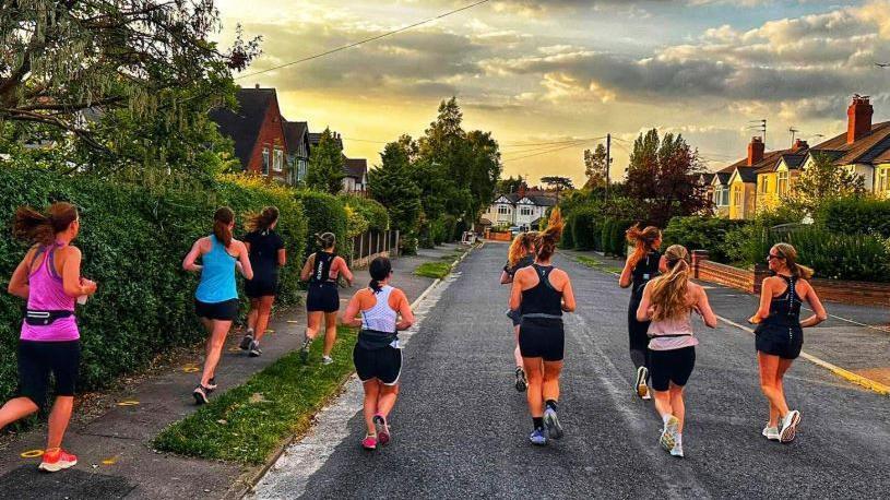 A group of women run along a residential road, which has houses on each side and grass verges. The women are running on the pavement and in the road, which is quiet.
