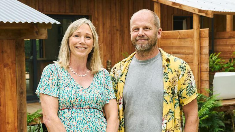 Marcus and Abigail standing in front of their home and smiling at the camera. Abigail's blonde hair sits on her shoulder, she wears a multi-coloured green dress with a white necklance. Marcus wears a yellow hawaian shirt with a grey t-shirt under it.