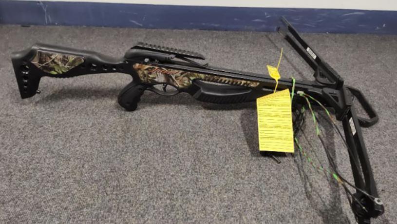 A black crossbow with camo trim sits on a carpeted floor. It has a yellow identification tag attached to it.
