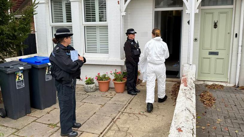 Two uniformed female police officers standing outside a white house with blinds in the windows. Another woman in white overalls is going into the house. 