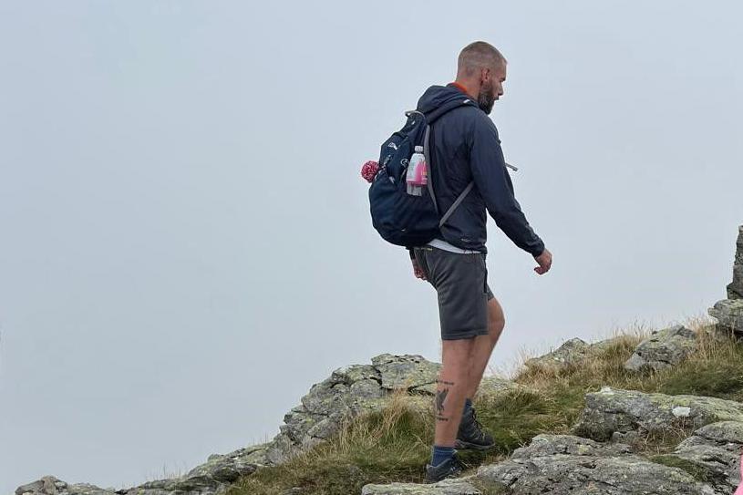 A side view of John Hall walking on a mountain in the Lake District. He has a back pack on and is wearing shorts. He has a tattoo on the back of his right calf. He has short cropped grey hair and facial hair. 