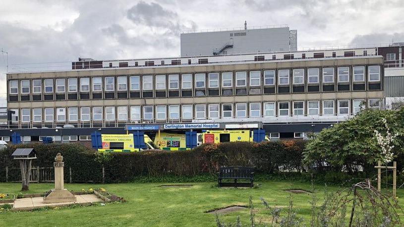 A long, wide building with numerous windows stands in the centre, with ambulances outside an entrance, a sign reads 'Darlington Memorial Hospital'. A park sits in front.