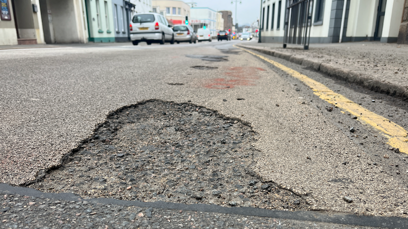 A pot hole on a main road with traffic on the left in the background.