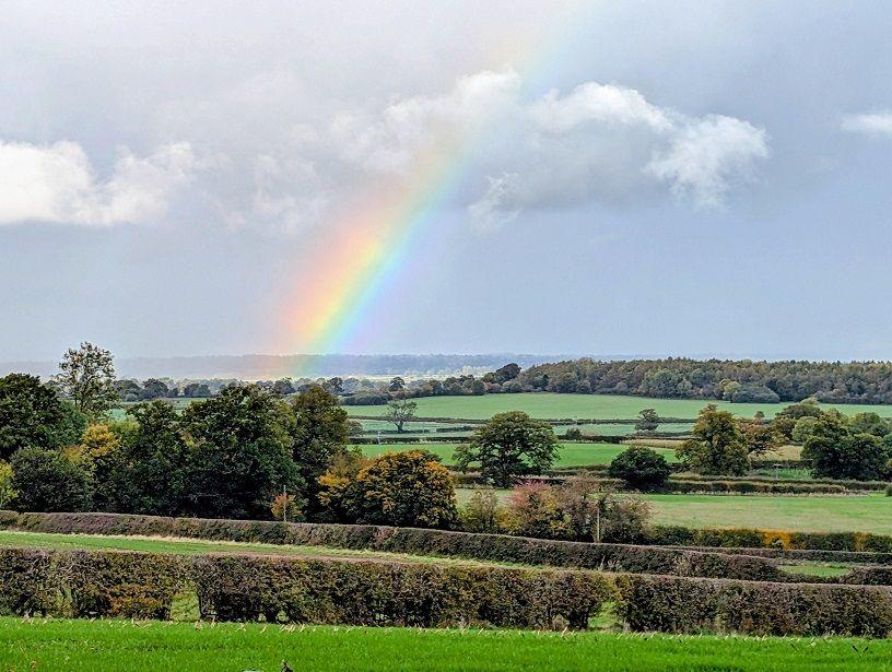 A rainbow is seen among the clouds over a rolling view of fields and hedges.