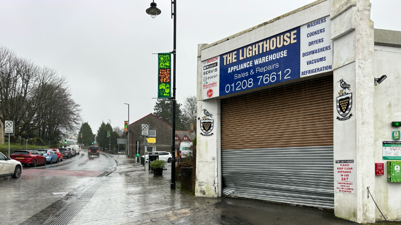 A street-view of a garage which has a dent in its metal barrier. There is a blue sign on top of the building with the words 'The Lighthouse' which is an appliance warehouse. There is a road to the left which has parked vehicles along the side of the road and a car driving in the distance.