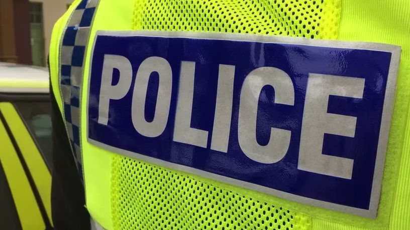 The back view of an officer's hi-vis yellow jacket. A blue sign on the back says police in white letters. A police car can be glimpsed on the left.