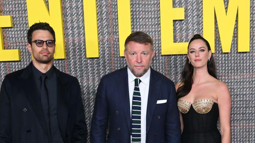 Theo James, Guy Ritchie and Kaya Scodelario pictured at the UK premiere of The Gentlemen in London in 2024. Guy Ritchie is dressed in a dark blue suit, white shirt and a tie featuring white stripes.