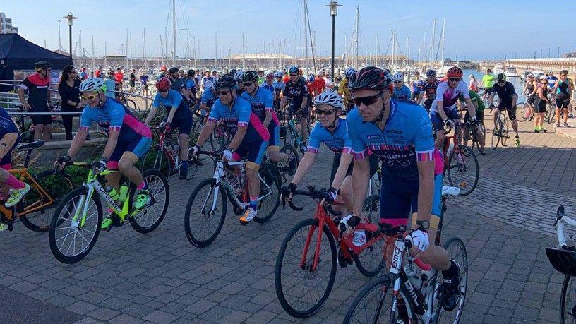 A picture of cyclists all on their bikes ready for a race. A majority of them are wearing a blue and pink outfit. The pier can be seen in the background.