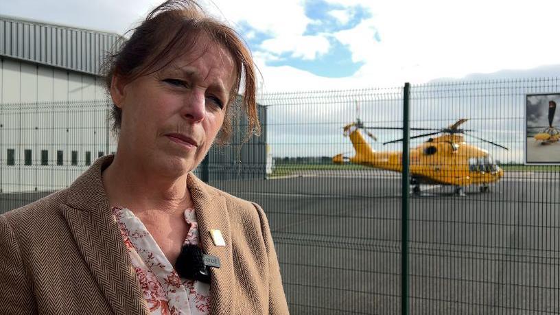 Karen Jobling is wearing a beige coat with a floral blouse. Her head is tilted to the right. She has brown hair. Behind her is a yellow helicopter.