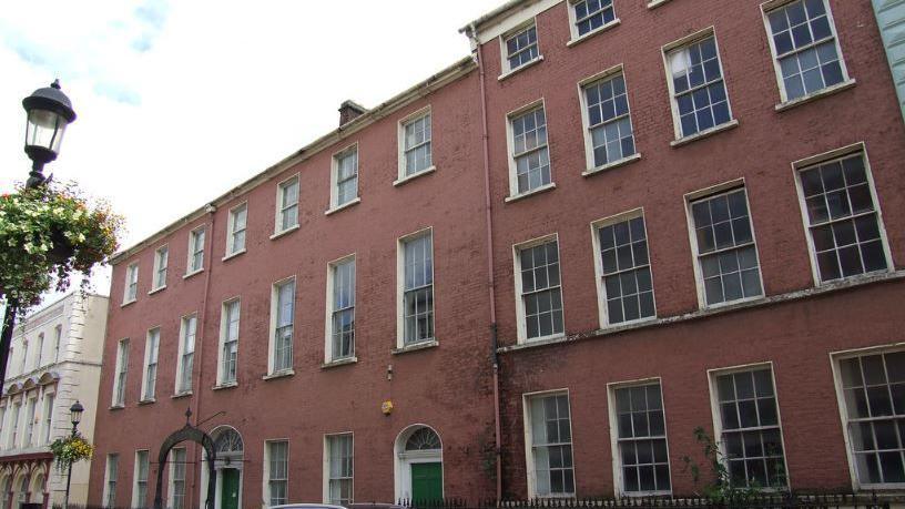 The former Convent of Mercy in Pump Street pictured in 2007.  The photo shows a row of red-brick Georgian terraced buildings with two green front doors.  The building on the left is a three storey, seven bay former hotel building and on the right is a four-storey, four bay former newspaper office, with a basement. 