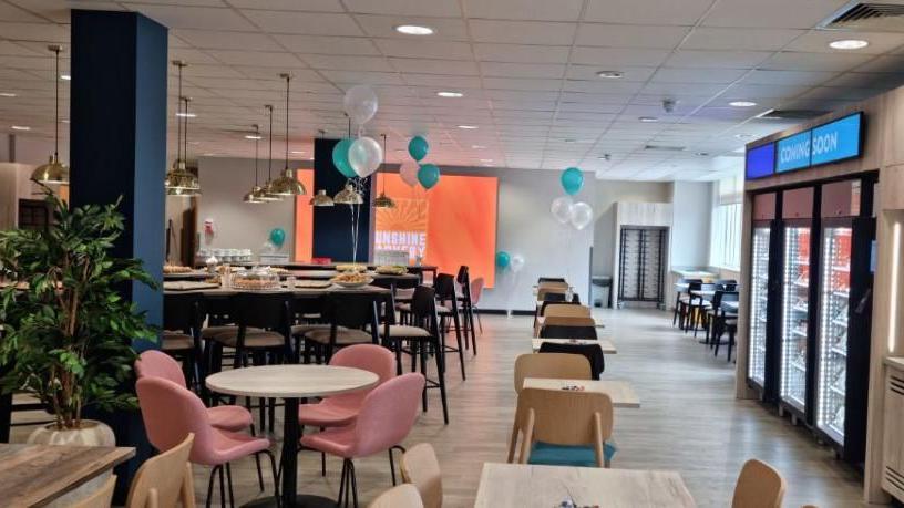 A cafe with pinks chairs and  white round tables, and decorated with green and white balloons. 