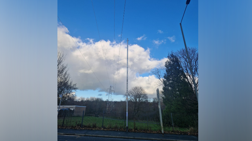 A high metal pole behind a fence, pointing towards blue sky, close to an electricity wire that is going overheard. 