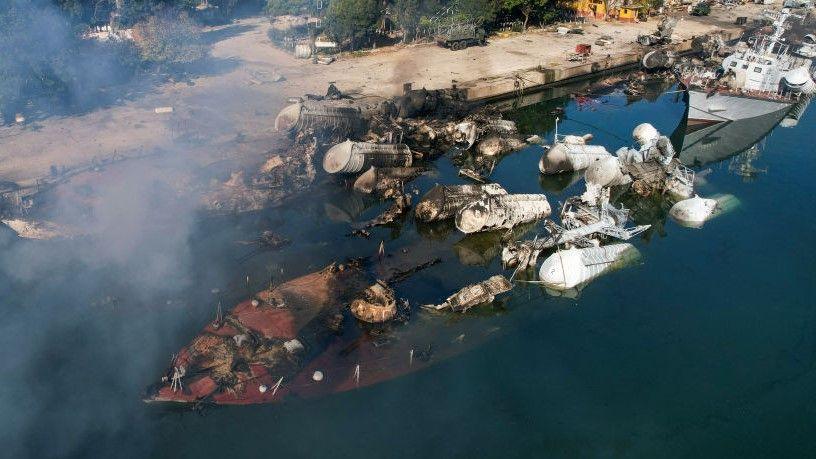 A sunken large boat close to the dockside in the Syrian port town of Latakia. There is smoke rising and partially submerged metallic structures. 