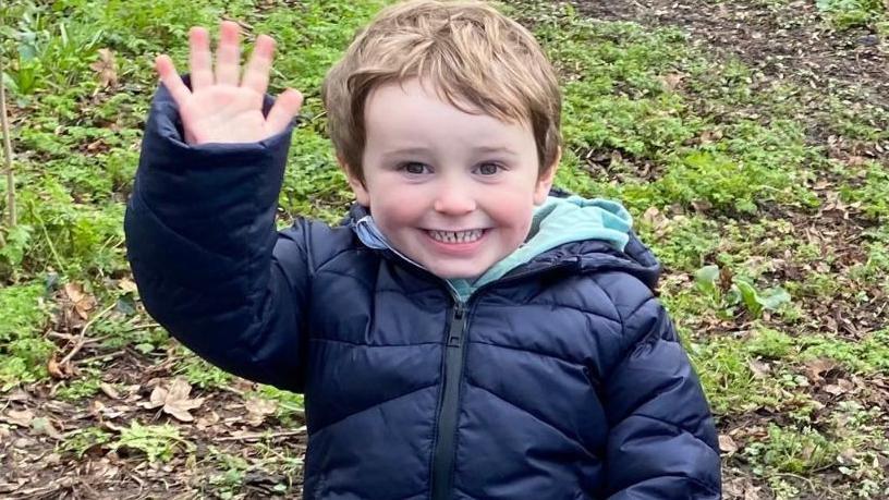 A young boy sitting of a log, smiling and waving. 