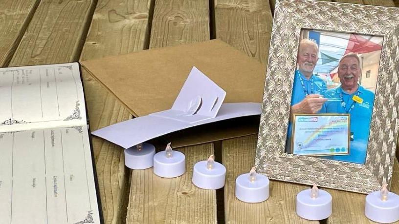 LED candles on a wooden table, alongside a gold photo frame. Inside the frame is a photo of two male volunteers wearing light blue shirts.