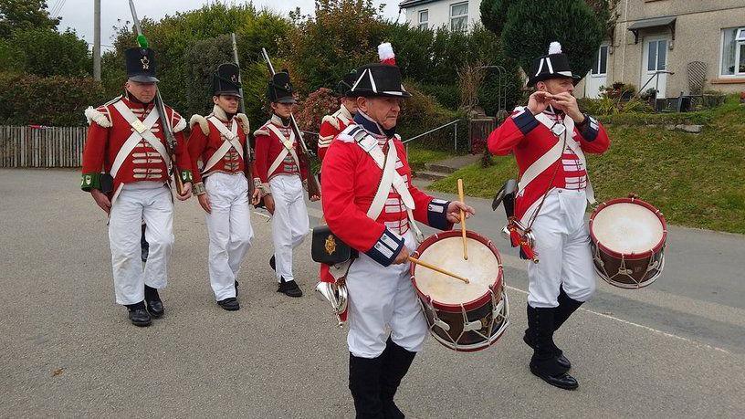 Members of the Plymouth Maritime Corps of Drums and members of the 32nd Cornwall Regiment of Foot, all in 1805 uniform, walk down the road.