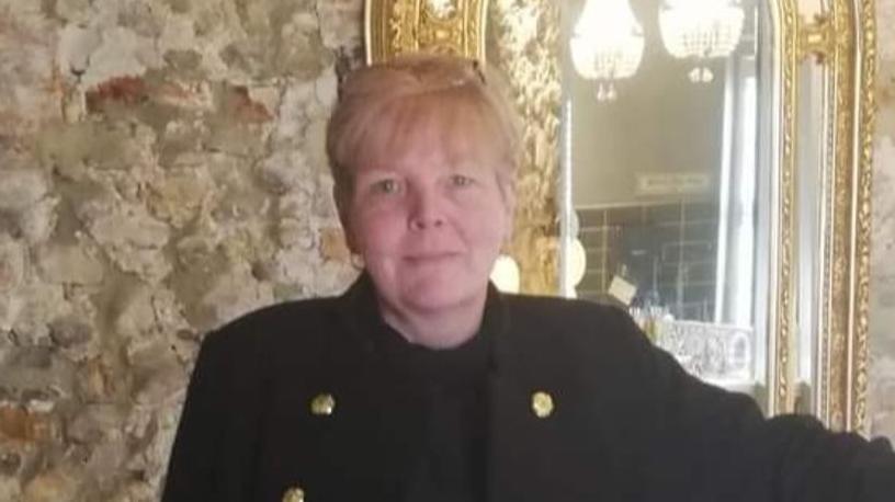 Head and shoulder shot of a woman with blonde hair and wearing a black coat looking straight at the camera. A mirror with a gilded border can be seen to her left