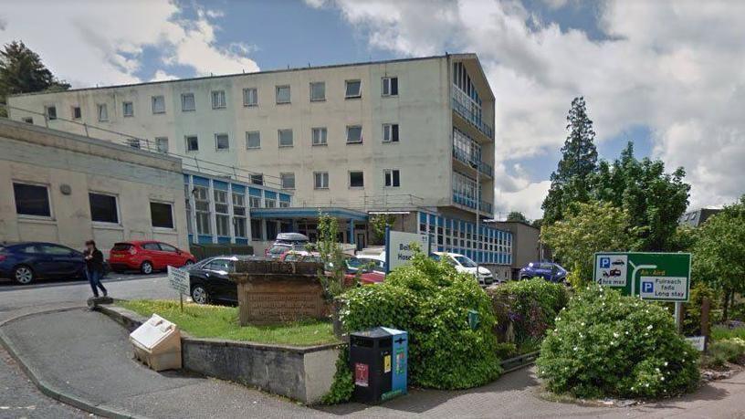 The hospital has multiple storeys and there is a car park with vehicles at the front of the building. There is also an area of trees and shrubs in front of the hospital.