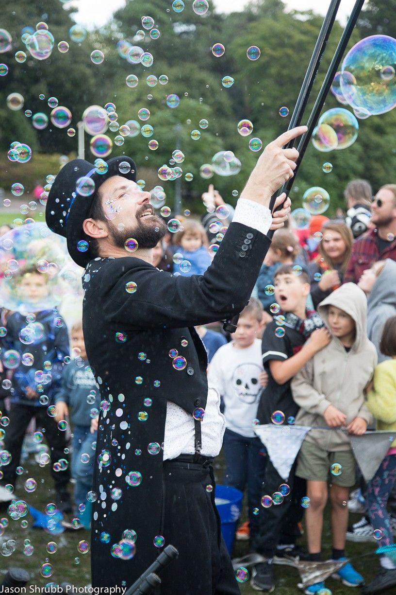 Man in a top hat juggles bubbles with a wand as a crowd watches on