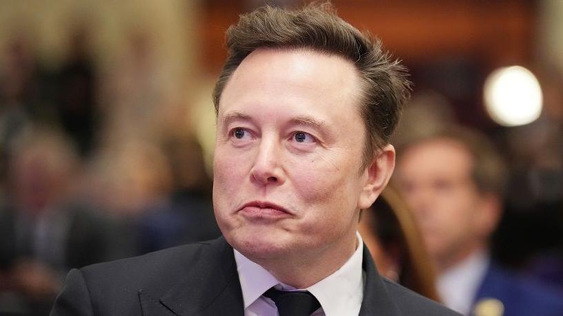 Elon Musk with a neutral expression wearing a suit and dark tie