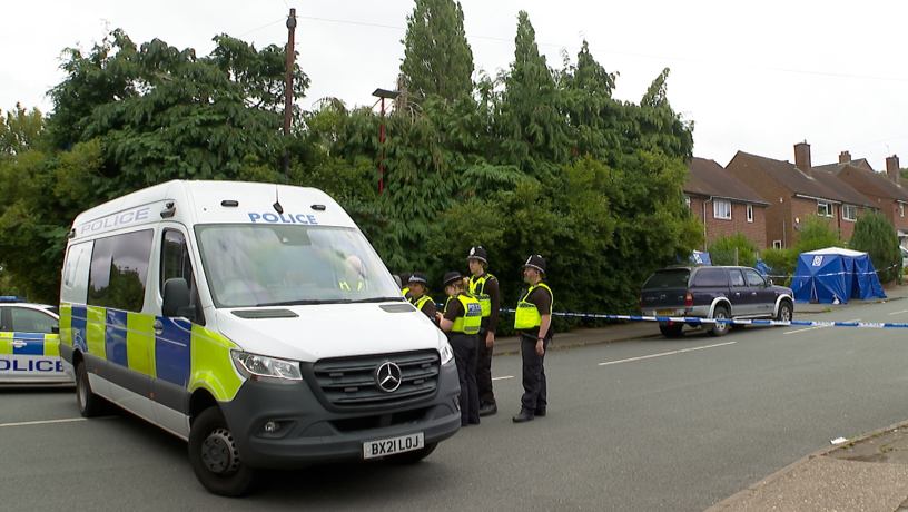 A police van and police car are parked across a road, with a number of officers to the side of the van. Behind them police tape is stretched over the road, with houses to the left side, one has blue tents outside