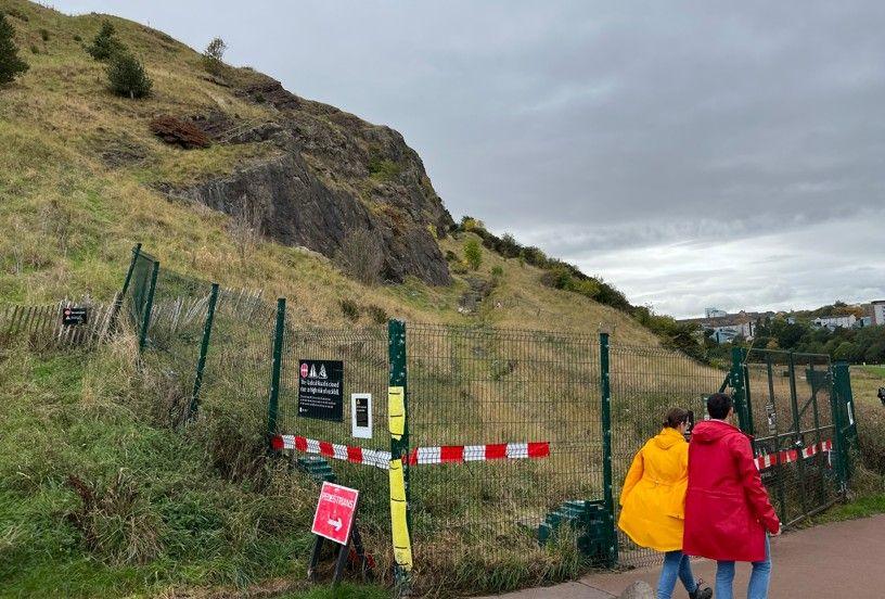 Two people in red and yellow jackets walk past a green fence that is keeping people out of the Radical Road Path in Holyrood Park. 