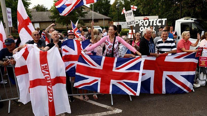 A group of people, some carrying large union jack and England flags. Some have placards with "Protect our..." and "Stop the boats" written on them. There are trees and a building behind them.