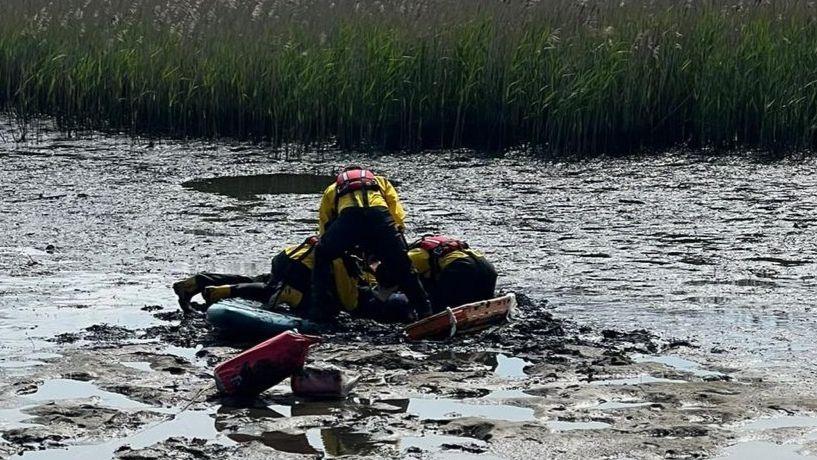 Mud with puddles on the surface with three RNLI crew members crouched together on the mud balanced on a stretcher to the right and a paddleboard to the left.