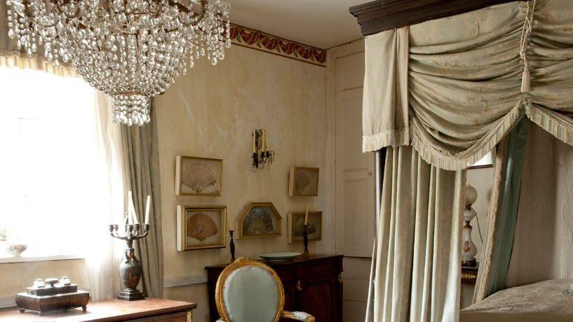 four poster bed with canopy. There is a chandelier hanging from ceiling, a wooden table and chair and paintings on the wall.