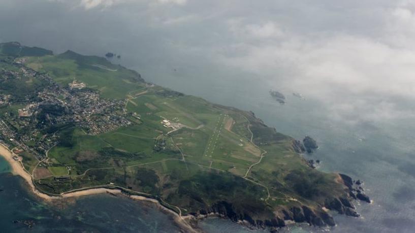 Drone image taken from above showing the island of Alderney and Alderney Airport. The island is mostly green with the main town of Saint Anne in the distance.