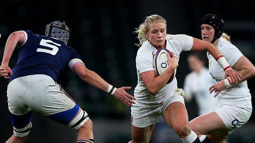 Three women playing rugby, one wears a blue shirt while the other two wear white. The woman in the middle is gripping a rugby ball in her right hand and is running out of reach of the woman wearing blue