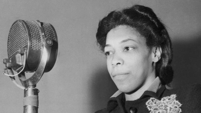 A black and white file image of Una Marson, a woman with dark pin-curled hair wearing a dark coloured jacket with flowers on, stands in front of a large old-fashioned silver microphone as she prepares to broadcast